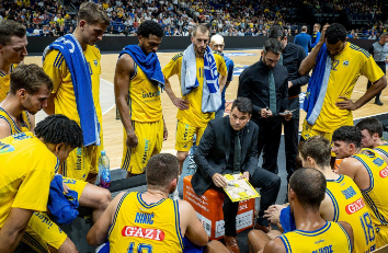 Foto: Am Spielfeldrand stehen die Spieler von ALBA Berlin um eine sitzende Person im Anzug herum.