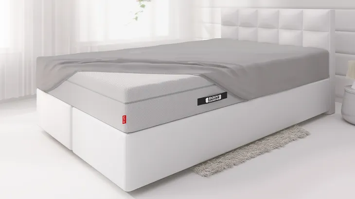In hellem Bett in hellem Schlafzimmer liegt die BODYGUARD Boxspring Matratze. Die Matratze ist bis auf eine Ecke bezogen mit dem BODYGUARD Spannbettlaken extra hoch.