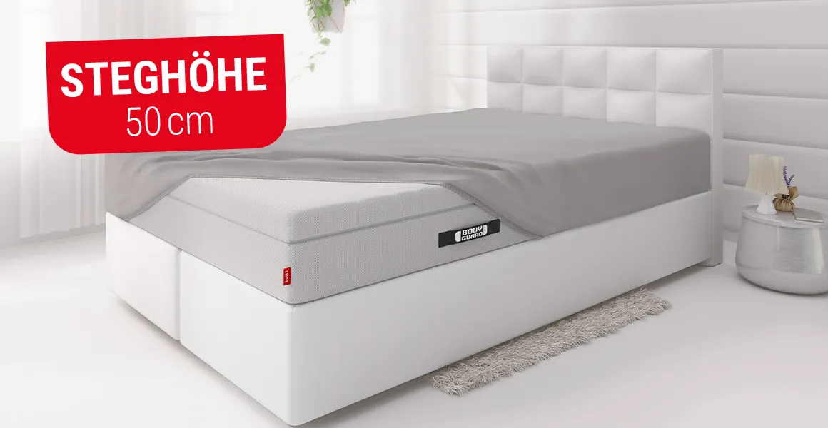 Die BODYGUARD Boxspring Matratze liegt in einem Bett und ist bis auf eine Ecke bezogen mit dem BODYGUARD Spannbettlaken extra hoch. Ein rot unterlegtes Feld mit Text: Steghöhe 50 cm.