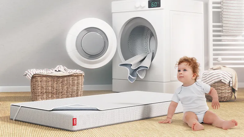 Die mit einem Nässeschutz bezogene BODYGUARD® KIndermatratze liegt vor einer geöffneten Waschmaschine, aus der eine weitere Nässeschutzauflage hängt.