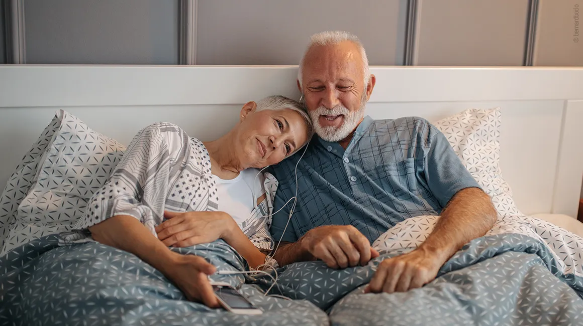 Foto: Zwei Personen sitzen zugedeckt im Bett und hören über Kopfhörer Musik. 