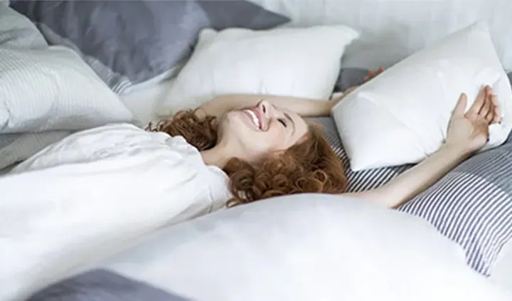 Eine Person liegt lächelnd und mit ausgestreckten Armen auf einem Bett voller Kissen.