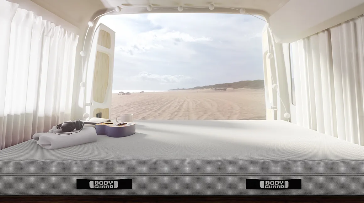 Das Innere eines Camping-Vans mit BODYGUARD Anti-Kartell-Matratze. Auf der Matratze liegen eine Ukulele sowie Strandausstattung. Durch die geöffneten Hintertüren ist ein Sandstrand zu sehen.