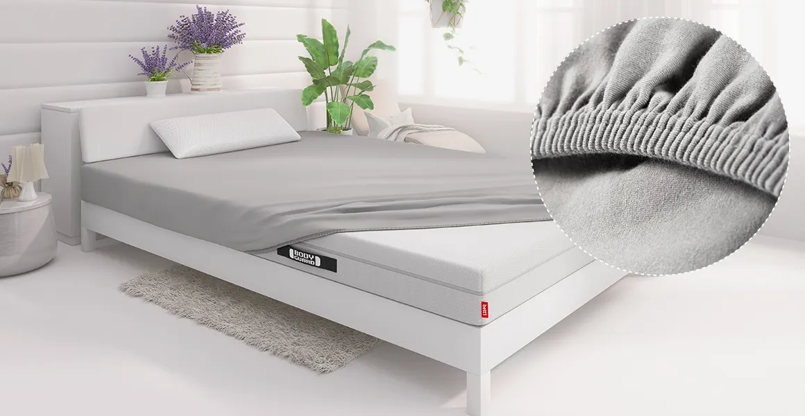 In einem Schlafzimmer liegt eine BODYGUARD Matratze in einem Bett, das mit dem Spannbettlaken 140x200 cm bezogen ist. Ein kreisförmiger Ausschnitt zeigt den Spanngummi in Nahaufnahme.