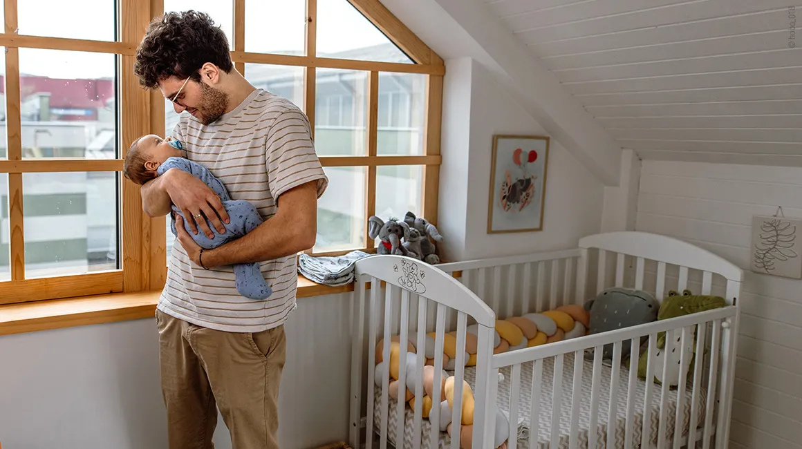 Foto: Eine Person steht neben einem Kinderbett und hat ein Baby auf dem Arm. 