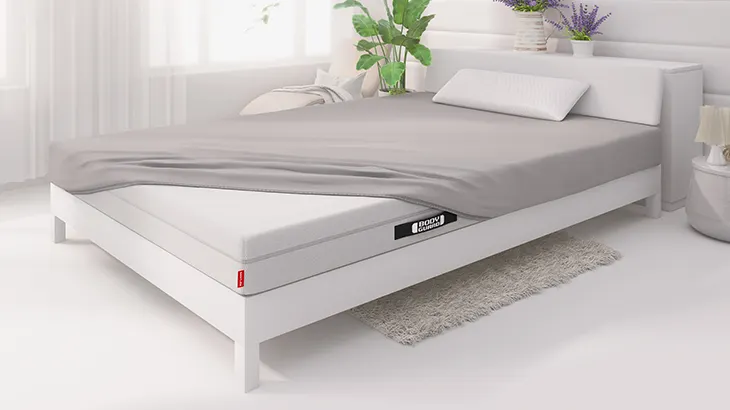 In einem hellen Schlafzimmer mit hellem Bett liegt die BODYGUARD Anti-Kartell-Matratze. Die Matratze ist bis auf eine Ecke bezogen mit dem BODYGUARD Spannbettlaken.