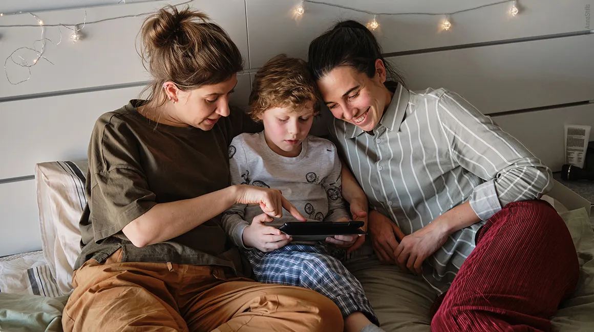 Foto: Zwei Personen und ein Kind liegen im Bett und schauen gemeinsam auf ein Tablet, das das Kind in den Händen hält. 