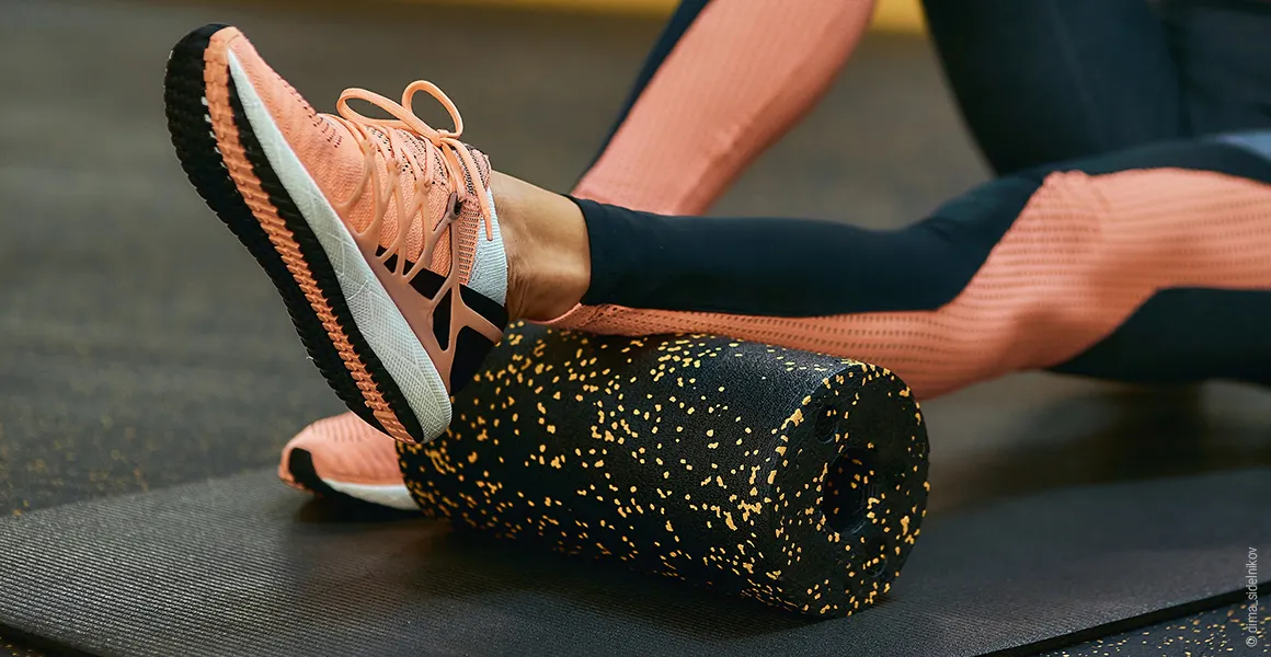 Foto: Eine Person in Sportkleidung rollt mit dem Unterschenkel über eine schwarze Faszienrolle auf einer Matte.
