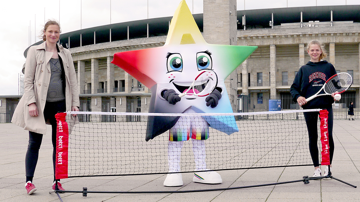 Foto: Zwei Personen und ein Maskottchen in Sternenkostüm stehen mit Tennischlägern hinter einem kleinen Tennisnetz vor dem Olympia Stadion.