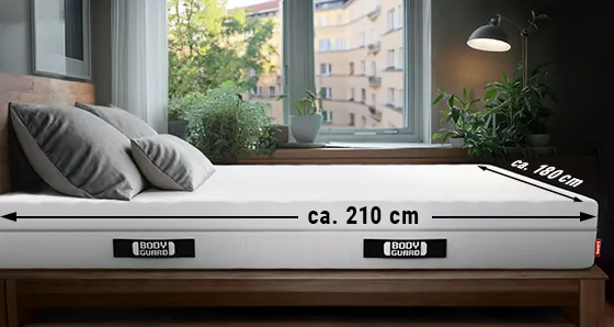 Eine BODYGUARD® Matratze liegt in einem Schlafzimmer. An der Längsseite ist ein Pfeil mit "ca. 210 cm", an der Breitseite ein Pfeil mit "ca. 180 cm" beschriftet.