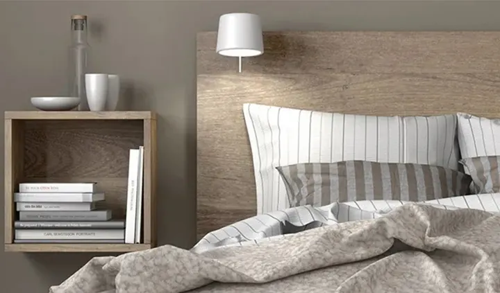 Ein Bett mit Kopfkissen und Decke. Am Kopfteil des Bettes hängt eine Leselampe, daneben ein Nachttisch.