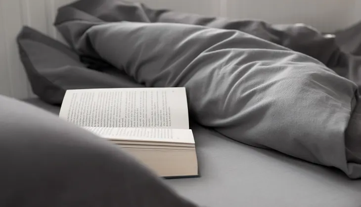Foto: Unter einer zurückgeklappten Bettdecke liegt ein aufgeschlagenes Buch auf einem Laken.
