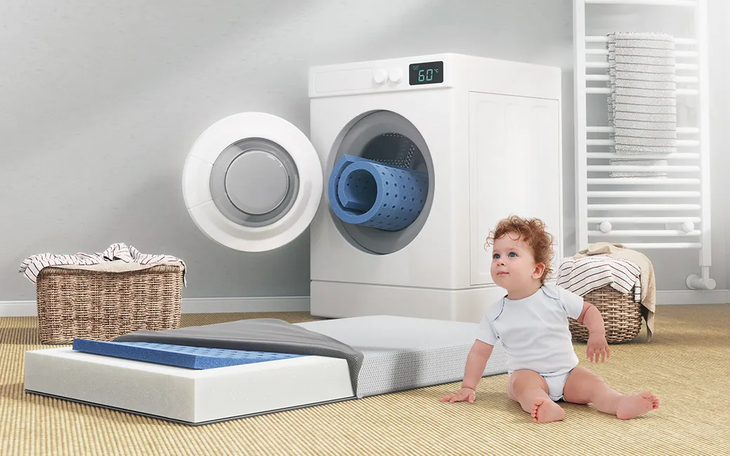 Die SuperBreeze Kindermatratze liegt mit halb abgezogenem Bezug und Blick auf die waschbare Liegefläche vor einer Waschmaschine, daneben sitzt ein Kleinkind.