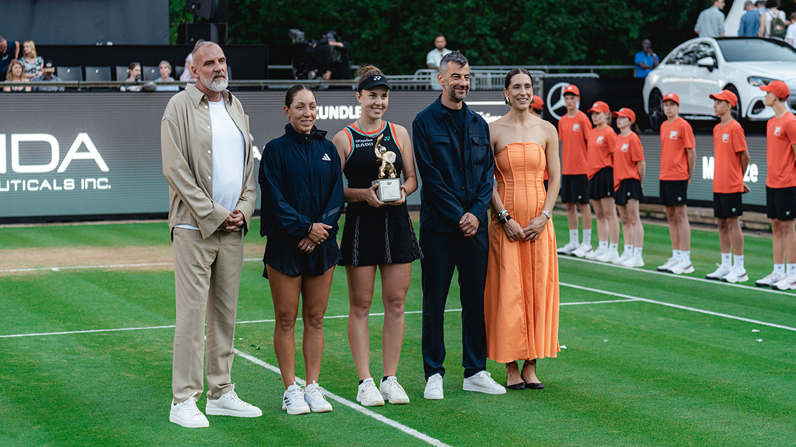 Fünf Personen stehen nebeneinander auf dem Tennisfeld. Die mittlere hält einen Pokal.
