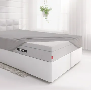 Die BODYGUARD® Boxspring Matratze ist bis auf eine Ecke mit dem Spannbettlaken extra hoch bezogen und liegt auf einem Boxspringbett in einem hellen Schlafzimmer.