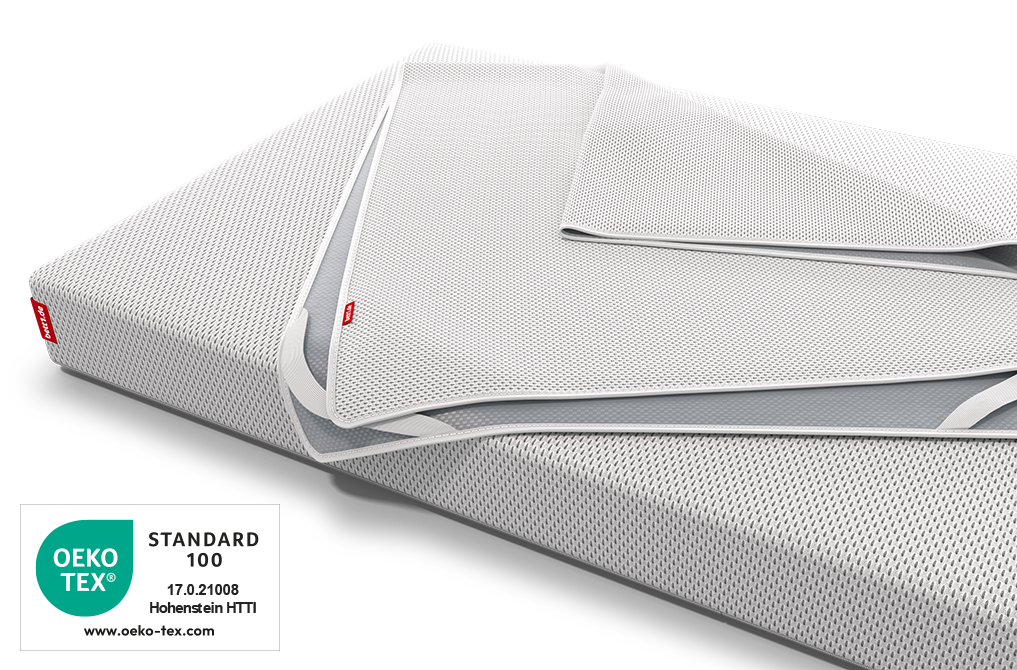 Der BODYGUARD® Nässeschutz für Kindermatratzen liegt zusammengelegt auf der bett1 Babymatratze, daneben das Oeko-Tex® Standard 100 Siegel.