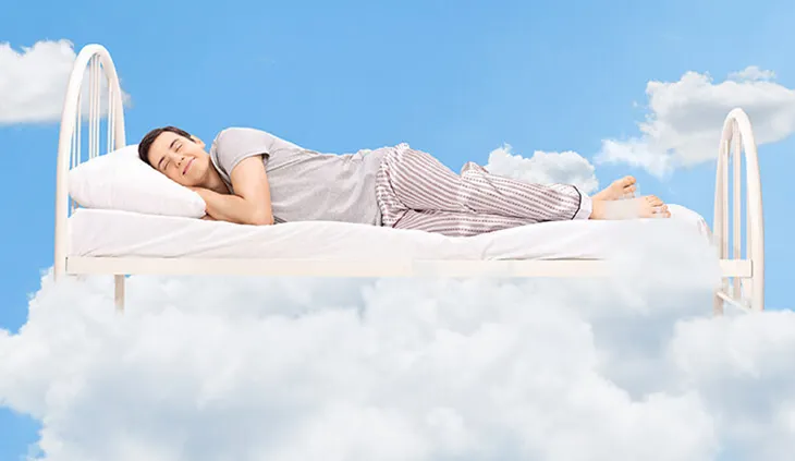 Eine Person liegt auf einem schwebenden Bett. Um das Bett herum sind Wolken, der Hintergrund ist himmelblau.