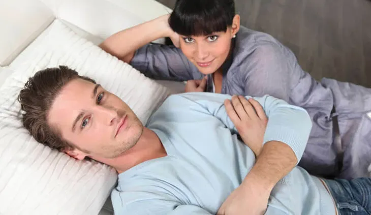 Zwei Personen liegen nebeneinander im Bett. Die eine rücklings und die Arme vor dem Oberkörper verschränkend. Die andere auf der Seite, mit dem Kopf abgestützt auf eine Hand.
