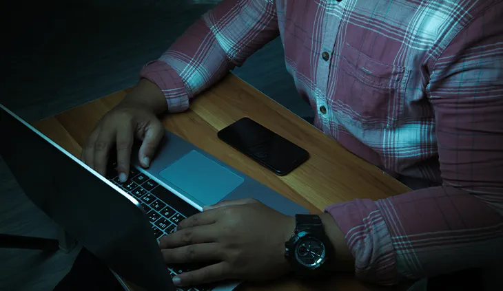 Einen Person sitzt bei schwummrigen Licht am Laptop. Vor ihr liegt außerdem ein Smartphone.