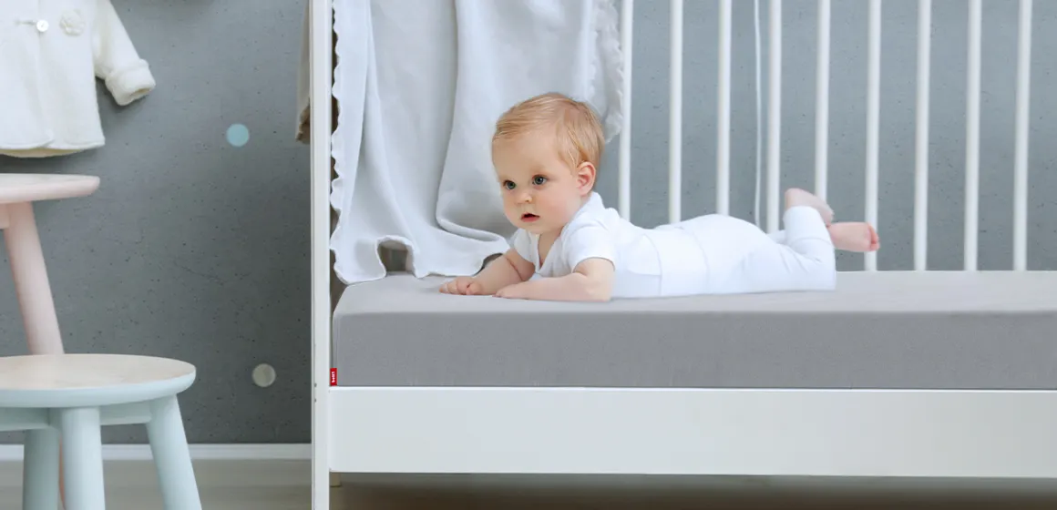 Ein Baby liegt im Kinderbett auf einer Babymatratze, die mit dem BODYGUARD Kinder-Spannbettlaken 70x140 cm in Silber bezogen ist.