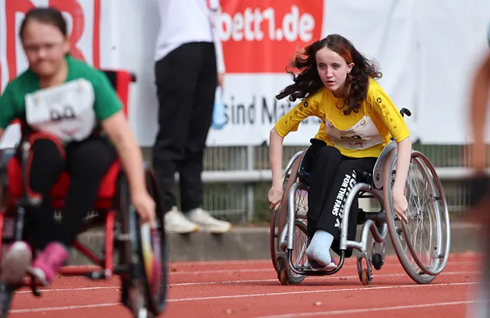 Einige Kinder in Rollstühlen mit angestrengt wirkenden Gesichtern und wehenden Haaren fahren ein Rennen.

