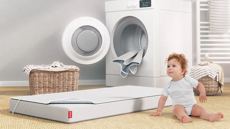 Die mit einem Nässeschutz bezogene BODYGUARD® KIndermatratze liegt vor einer geöffneten Waschmaschine, aus der eine weitere Nässeschutzauflage hängt.