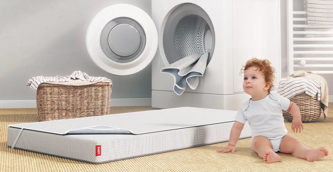 In einem Badezimmer sitzt ein Kleinkind vor einer Waschmaschine. Daneben liegt eine SuperBreeze Kindermatratze auf dem Boden. Sie ist mit dem BODYGUARD Nässeschutz bezogen.