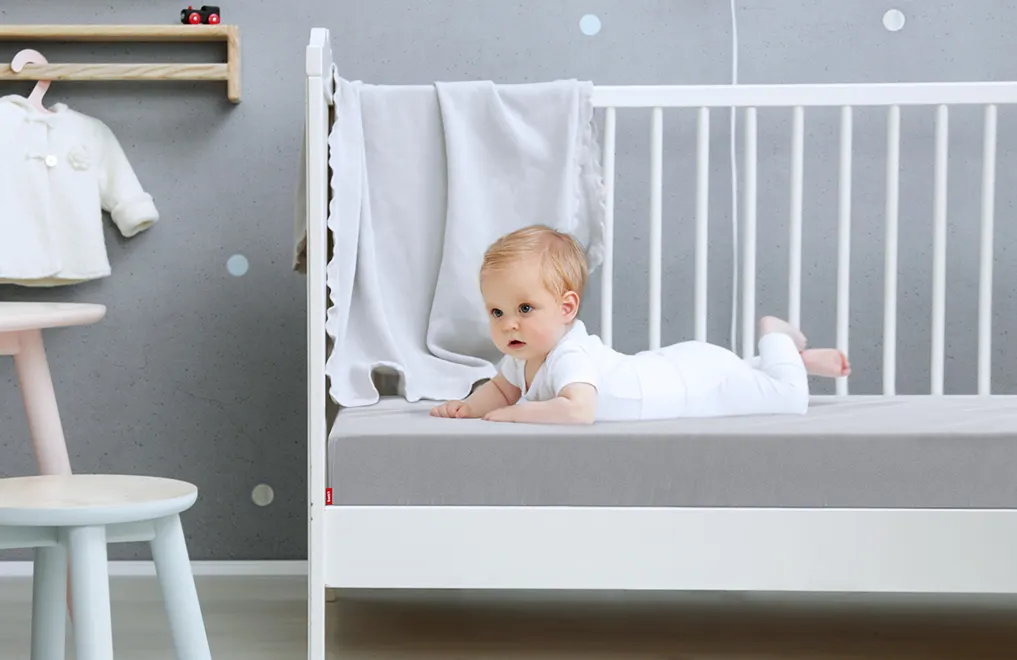 Ein Baby liegt in einem Kinderbett auf einer Babymatratze, die mit dem Kinder-Spannbettlaken in Silber bezogen ist.	