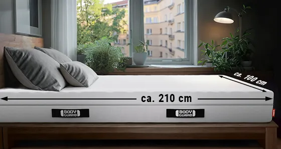 Eine BODYGUARD® Matratze liegt in einem Schlafzimmer. An der Längsseite ist ein Pfeil mit "ca. 210 cm", an der Breitseite ein Pfeil mit "ca. 100 cm" beschriftet.