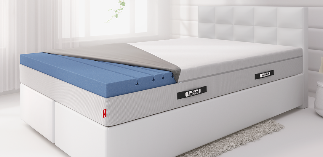 Foto: Bett mit einer BODYGUARD Boxspring Matratze 180x220 cm in einem hellen Schlafzimmer. Der Matratzenbezug ist halb zurückgeschlagen, sodass der obere Teil des zweiteiligen Matratzenkerns zu sehen ist