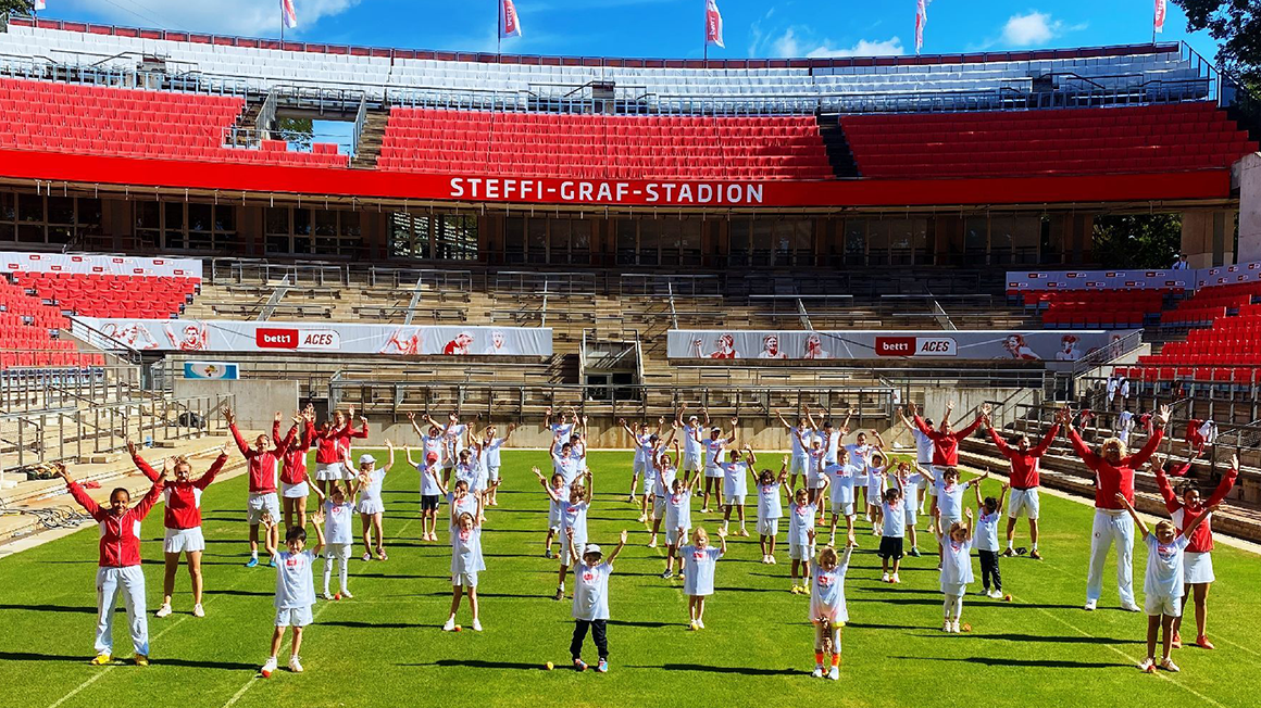 Vor einer roten Tribüne stehen Kinder und Erwachsene in roter und weißer Sportbekleidung in Reihen und heben beide Arme in die Luft.