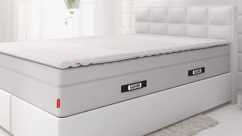 Auf einem weißen Boxspringbett liegt die extra hohe BODYGUARD Matratze und darauf der Topper 140x200 cm.