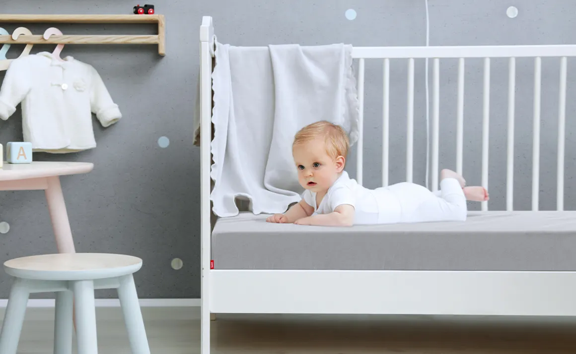 Ein Baby liegt in einem Kinderbett auf einer Babymatratze, die mit dem Kinder-Spannbettlaken in Silber bezogen ist.	