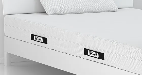 Auf einem einfachen Holzbett liegt die BODYGUARD Matratze 100x220 cm mit Kissen.