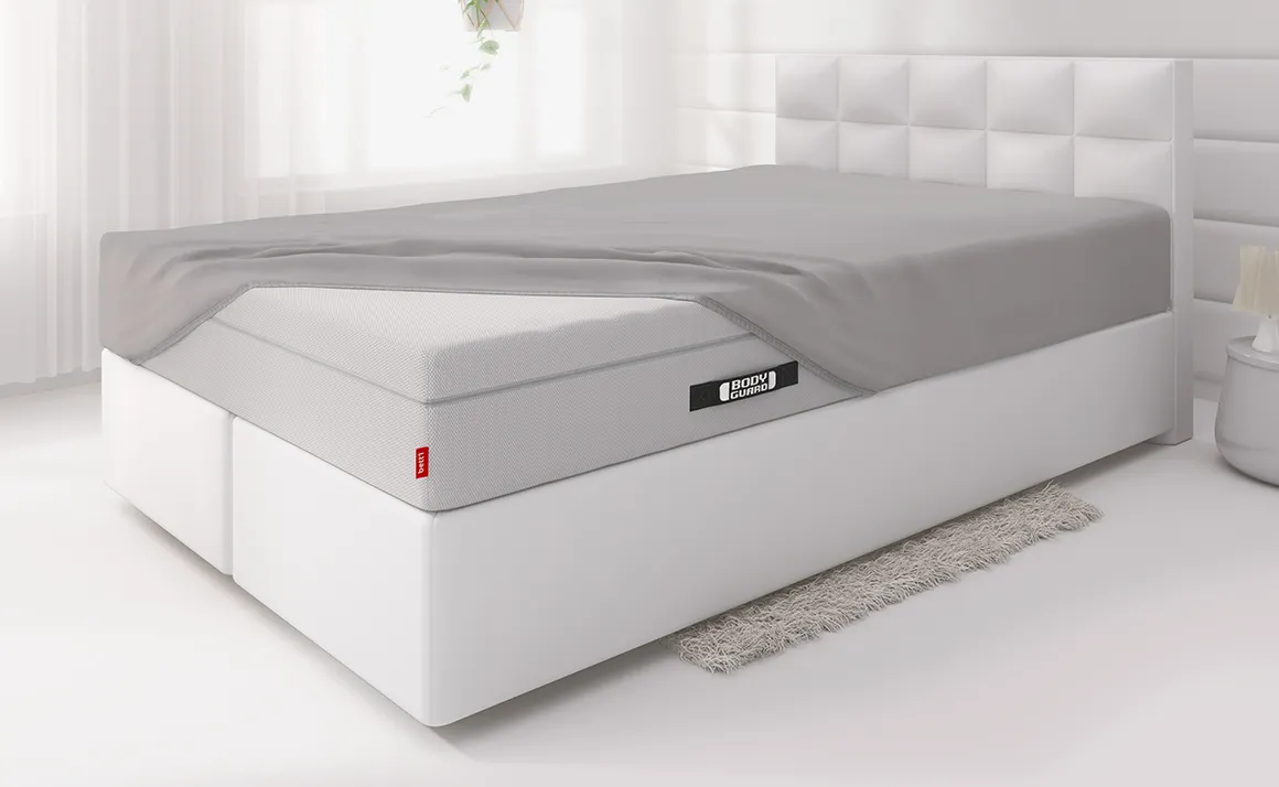 Die BODYGUARD® Boxspring Matratze ist bis auf eine Ecke mit dem Spannbettlaken extra bezogen und liegt auf einem Boxspringbett in einem hellen Schlafzimmer	