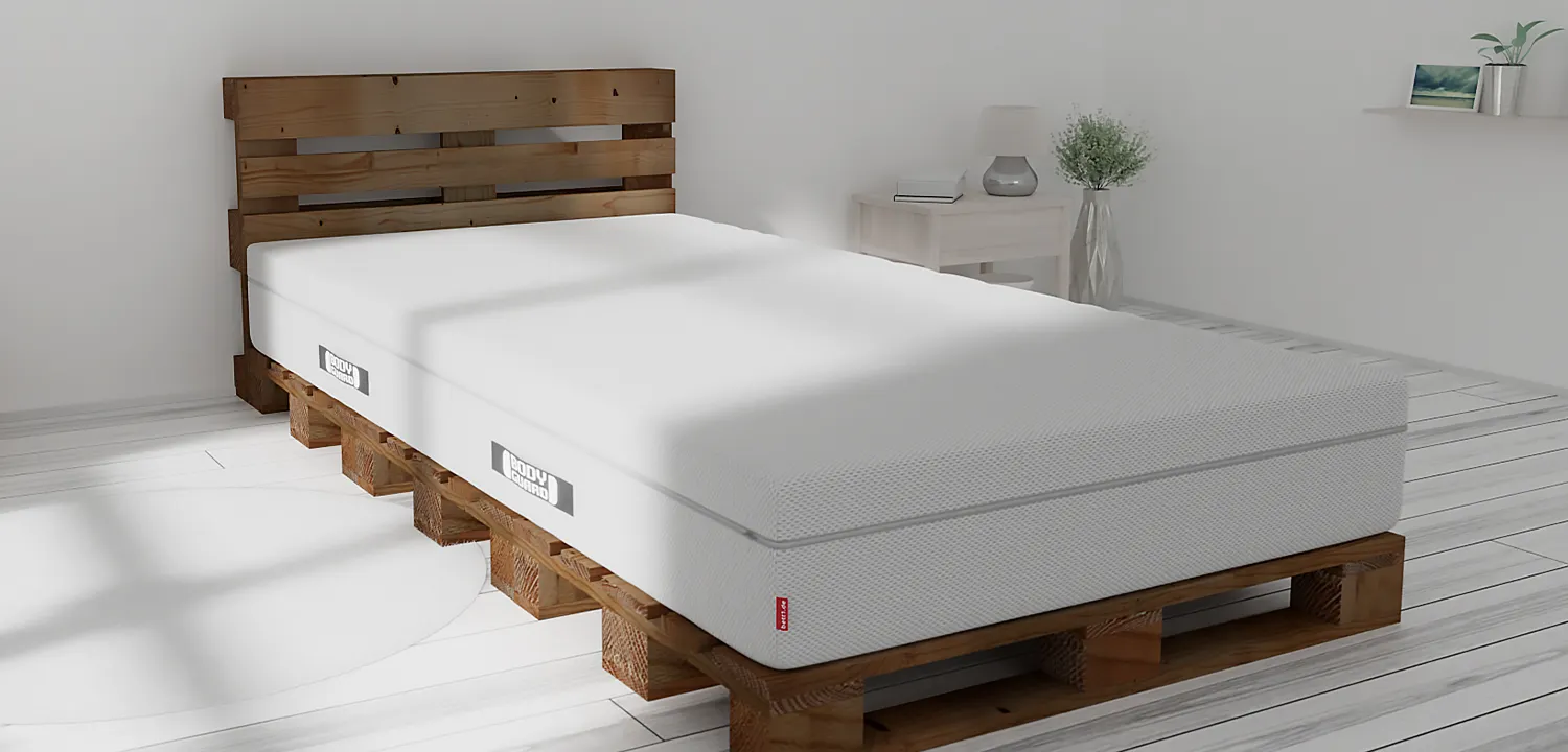 In einem hellen Schlafzimmer steht ein Palettenbett aus dunklem Holz, darauf die BODYGUARD Boxspring Matratze 100x220 