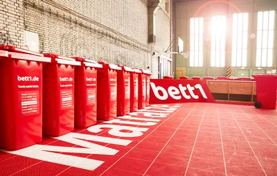 Foto: Eine Reihe von roten bett1-Tonnen auf einem roten Hallentennisplatz.