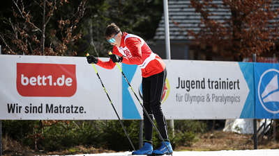 Vor einer Werbebande steht eine Person vornübergebeugt auf Skiern und sticht Ski-Stöcke in den Boden.