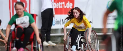 Einige Kinder in Rollstühlen mit angestrengt wirkenden Gesichtern und wehenden Haaren fahren ein Rennen.