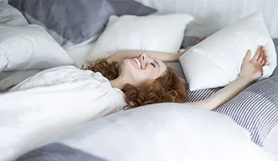 Eine Person liegt lächelnd und mit ausgestreckten Armen auf einem Bett voller Kissen.