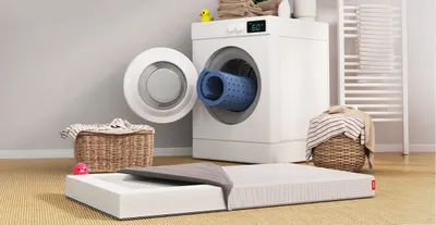 Die SuperBreeze Kindermatratze mit halb geöffnetem Bezug liegt vor einer Waschmaschine. Eine waschbare Liegefläche schaut zusammengerollt aus der Waschmaschine.