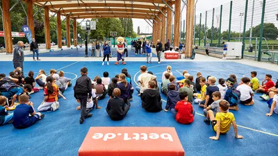 Zahlreiche Kinder sitzen auf einem Sportplatz und blicken in Richtung des Maskottchens vom bett1.de Multisportevent.
