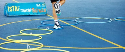 Auf einem Spielfeld liegt ein Parkour aus Hoola-Hoop-Reifen und Kinderbeine springen hindurch, dahinter ein Banner mit Text: ISTAF macht Schule.