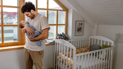 Foto: Eine Person steht neben einem Kinderbett und hat ein Baby auf dem Arm. 