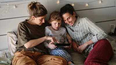 Foto: Zwei Personen und ein Kind liegen im Bett und schauen gemeinsam auf ein Tablet, das das Kind in den Händen hält. 