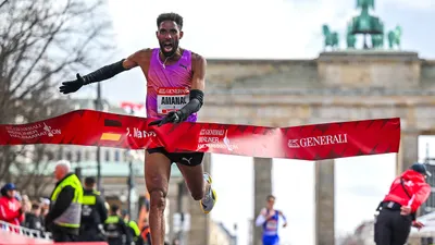 The home straight of the Berlin Generali Half Marathon at the Brandenburg Gate. A runner sprints through the finish line banner.
